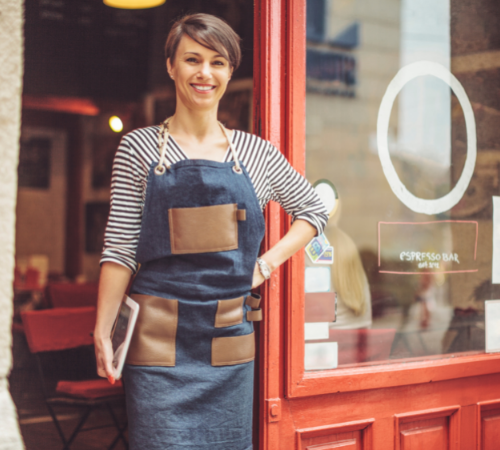 Woman in apron happily standing outside of shop and holding a tablet 