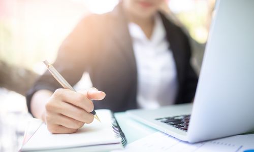 Person writing in a note pad with an open laptop